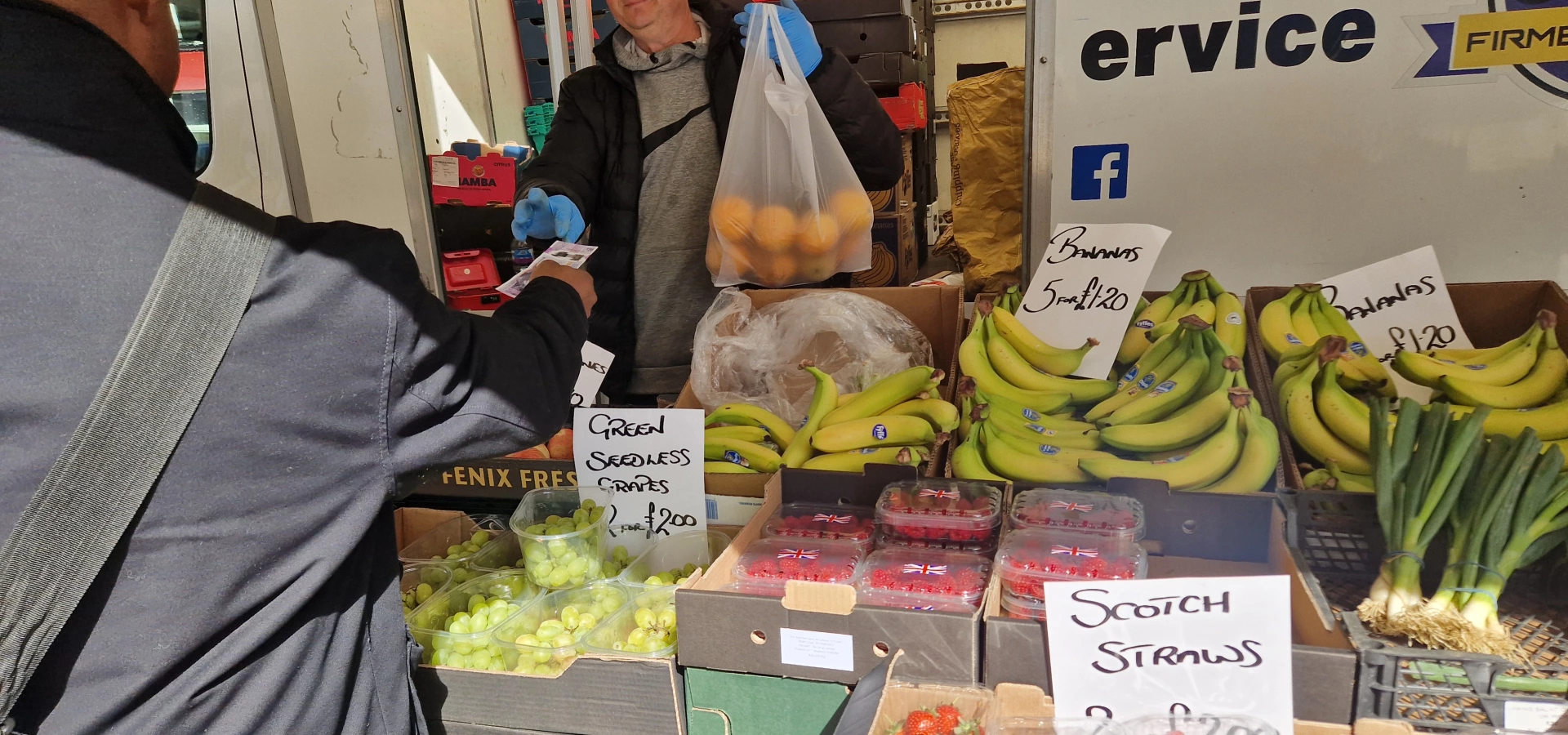 A man paying a fruit stall holder at a local market. There are bananas, grapes, raspberries and strawberries stacked up on the table.